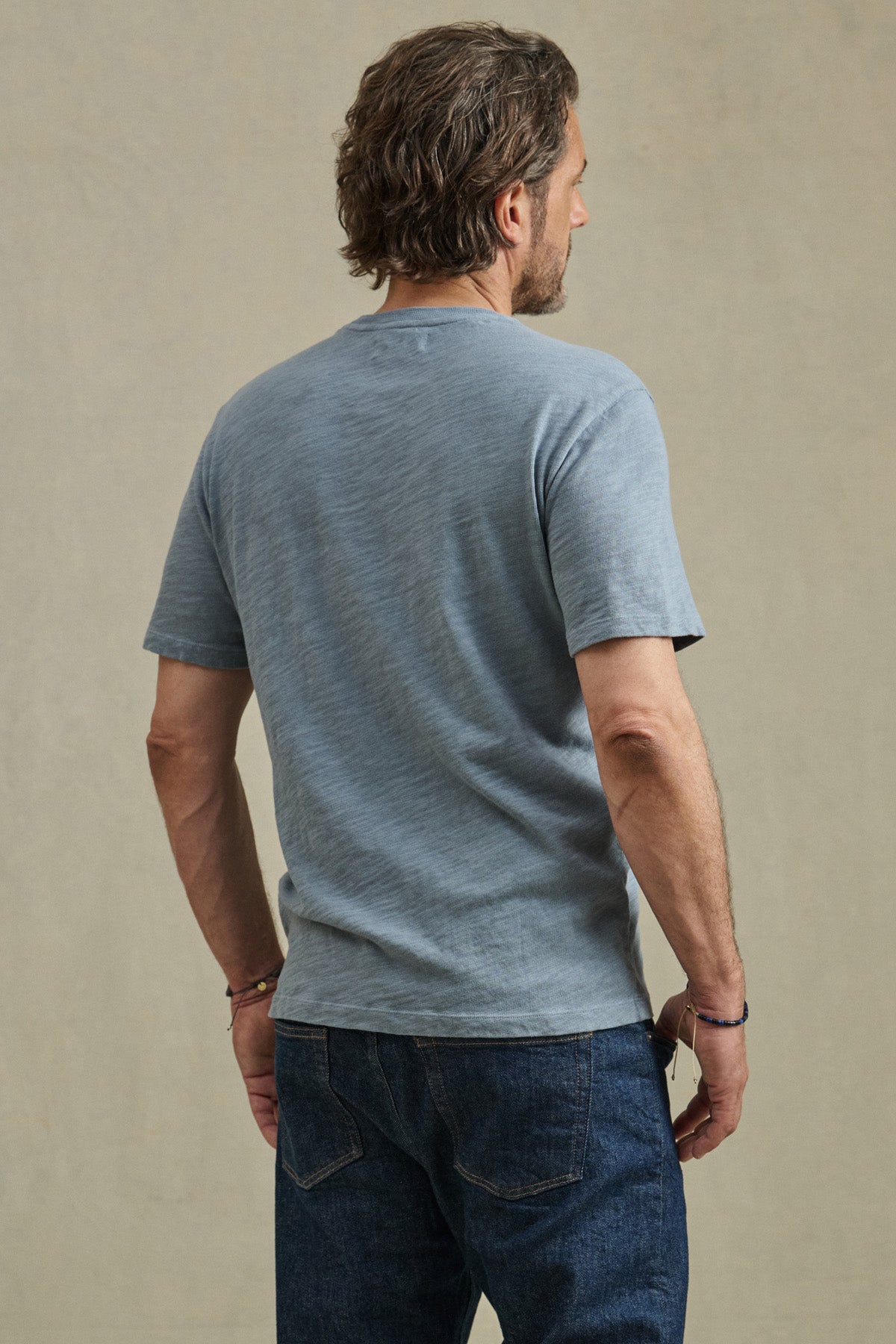 A man with wavy brown hair stands facing away from the camera against a plain background, wearing the PAUL TEE by Velvet by Graham & Spencer in light blue cotton slub and dark blue jeans.