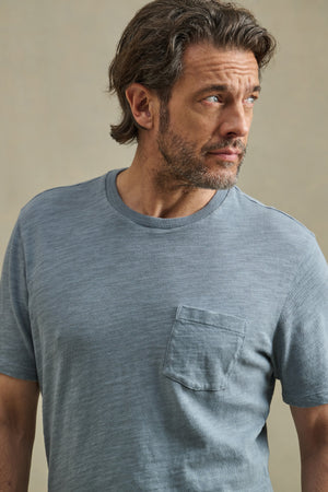 Man with medium-length brown hair and beard wears the PAUL TEE by Velvet by Graham & Spencer, a light blue crew neck cotton slub shirt with a chest pocket, as he looks to his left against a neutral background.