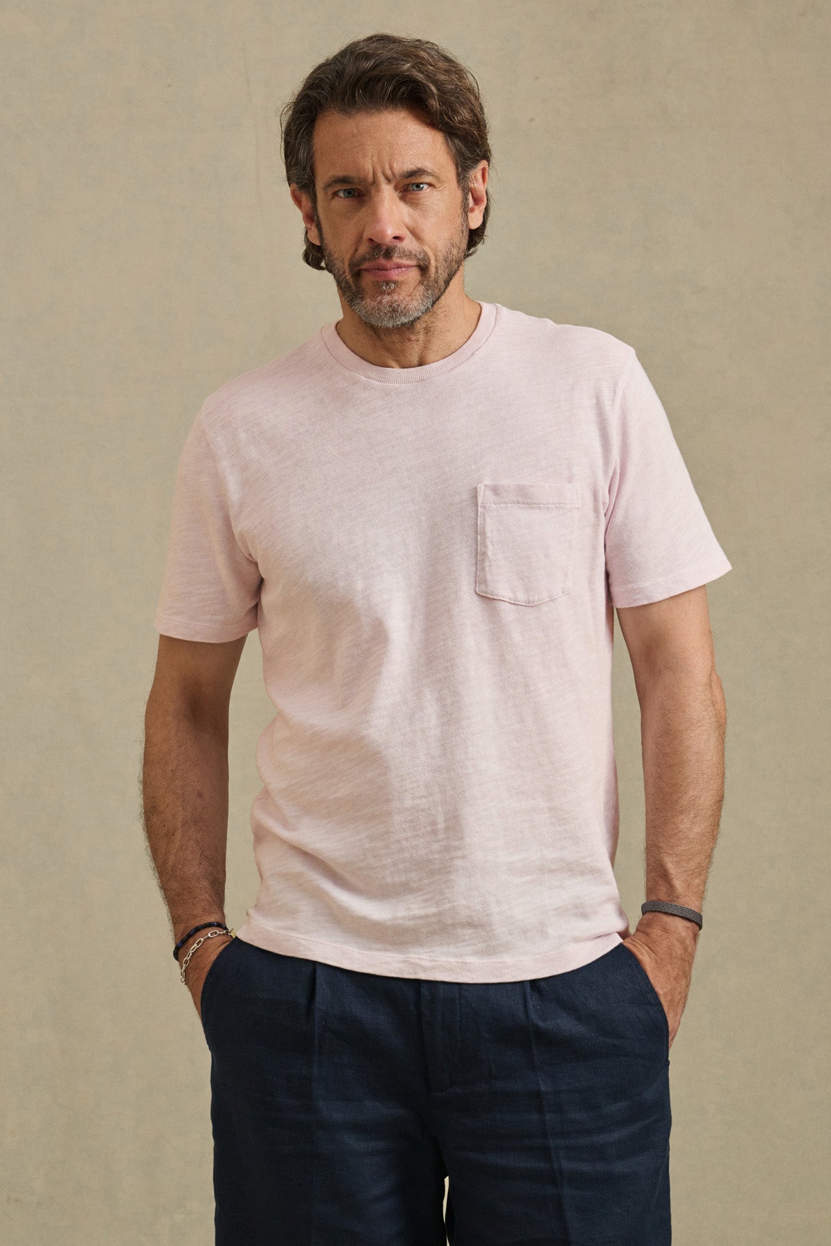 A man with wavy hair and a beard wears the Velvet by Graham & Spencer PAUL TEE—a light pink cotton slub crew neck—paired with dark pants, standing against a plain beige background with his hands in his pockets.