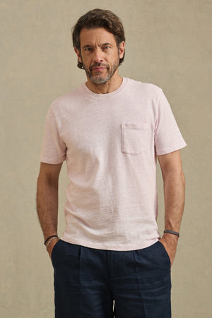 A man with wavy hair and a beard wears the Velvet by Graham & Spencer PAUL TEE—a light pink cotton slub crew neck—paired with dark pants, standing against a plain beige background with his hands in his pockets.