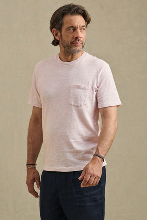 A man with brown hair and a beard wears the PAUL TEE by Velvet by Graham & Spencer, a pale pink cotton slub crew neck shirt with a chest pocket, styled with dark pants against a plain background.