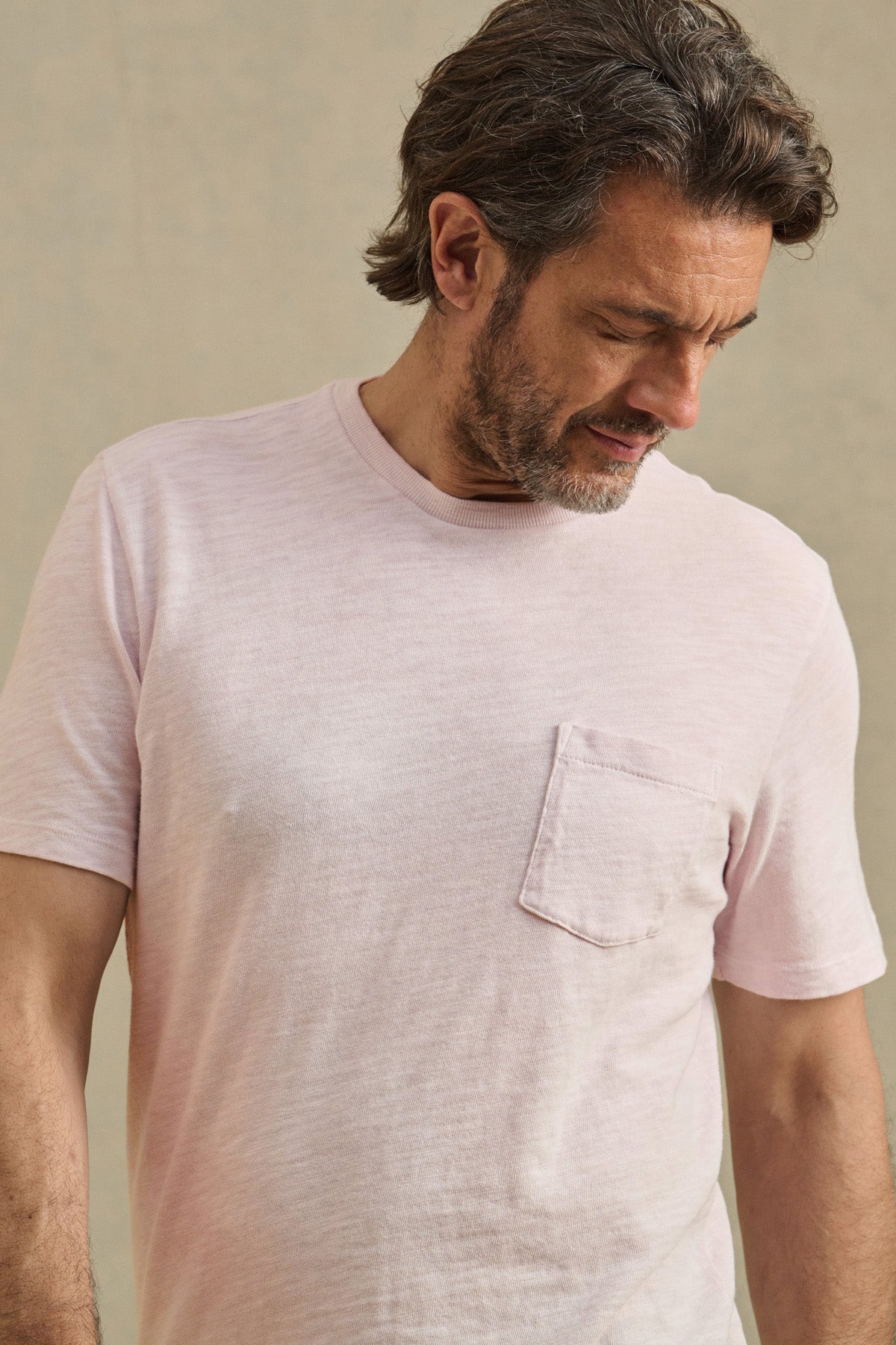 A man with short brown hair and a beard wears the PAUL TEE by Velvet by Graham & Spencer, a light pink 100% cotton crew neck with a chest pocket, looking down against a neutral background.-41491030900929
