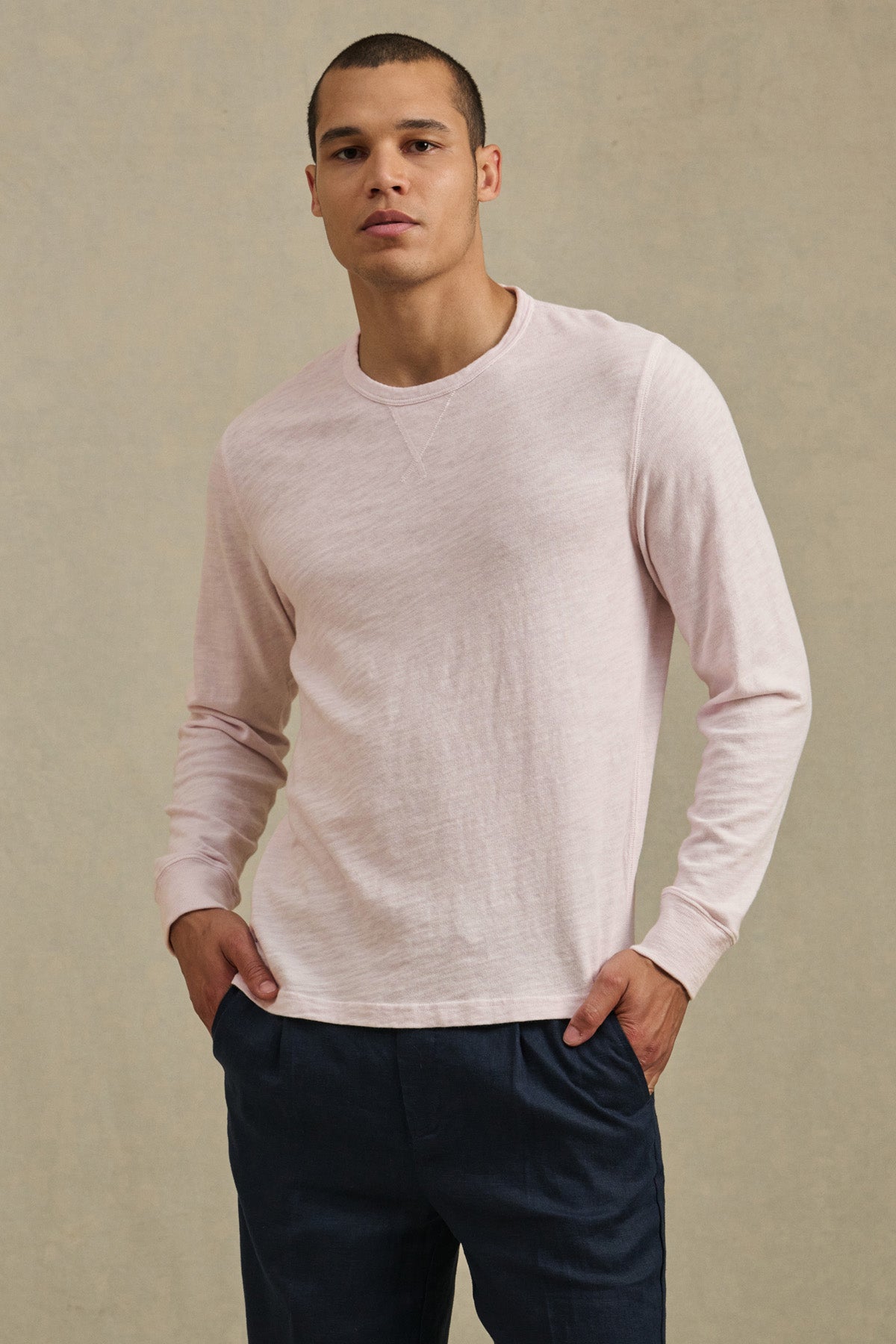 A person wears the ZANE TEE in light pink by Velvet by Graham & Spencer with dark blue pants, standing against a plain, light background with hands in pockets.-41480104345793