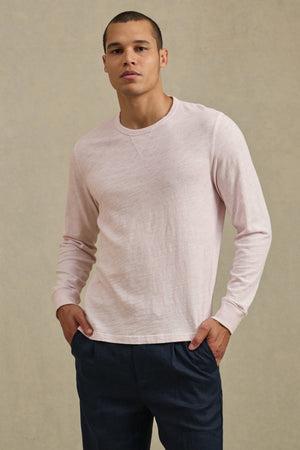 A person wears the ZANE TEE in light pink by Velvet by Graham & Spencer with dark blue pants, standing against a plain, light background with hands in pockets.