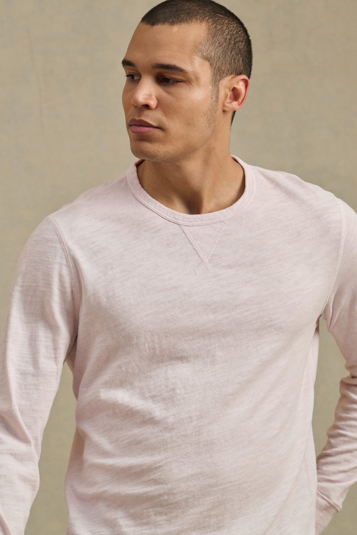 A man wearing the ZANE TEE by Velvet by Graham & Spencer, a light pink cotton slub long sleeve shirt, stands against a plain beige background and looks to his left.-41480104411329