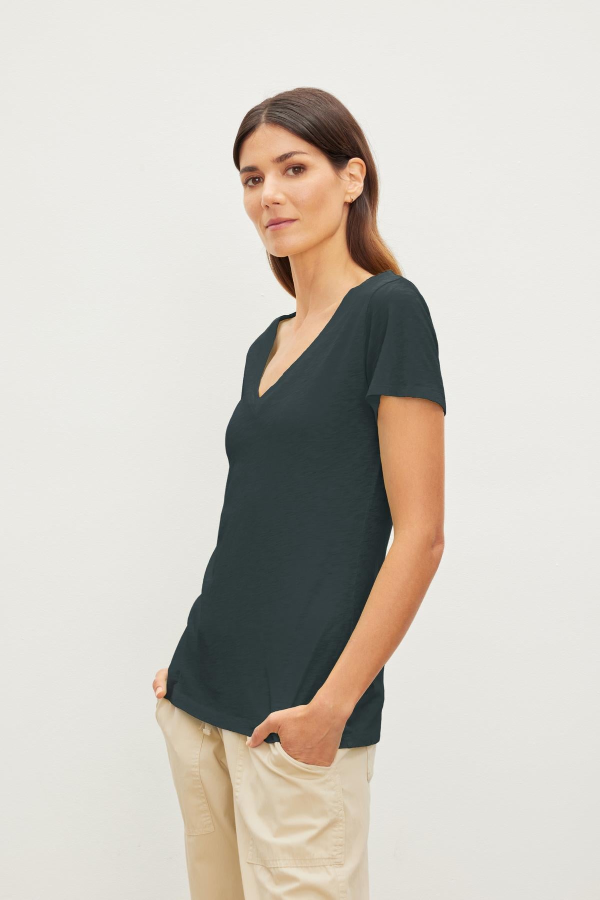   A woman with long brown hair wearing a dark green LILITH TEE by Velvet by Graham & Spencer and light beige pants stands against a plain white background, exuding an air of luxurious softness. 