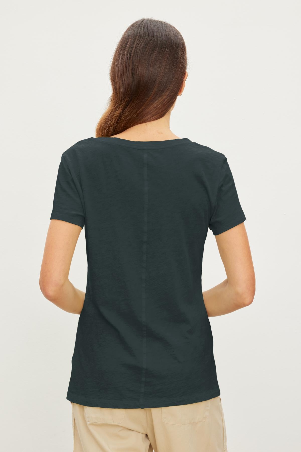 A person with long hair is shown from the back, wearing a dark green short-sleeved V-neck LILITH TEE by Velvet by Graham & Spencer and beige pants, standing against a plain white background. The shirt's cotton slub fabric adds a touch of luxurious softness.-37241138741441