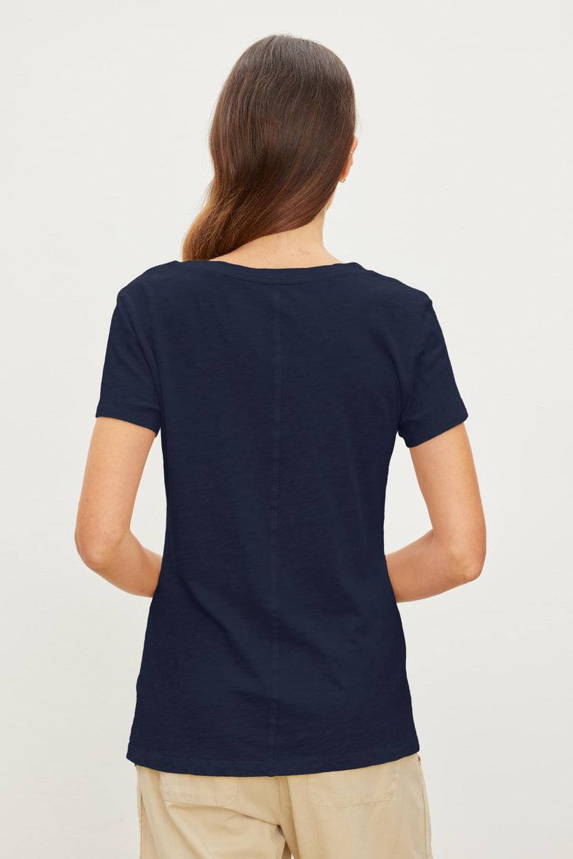 A person with long hair, seen from behind, is wearing a navy blue short-sleeve LILITH TEE by Velvet by Graham & Spencer in a flattering fit and light-colored pants against a plain background.