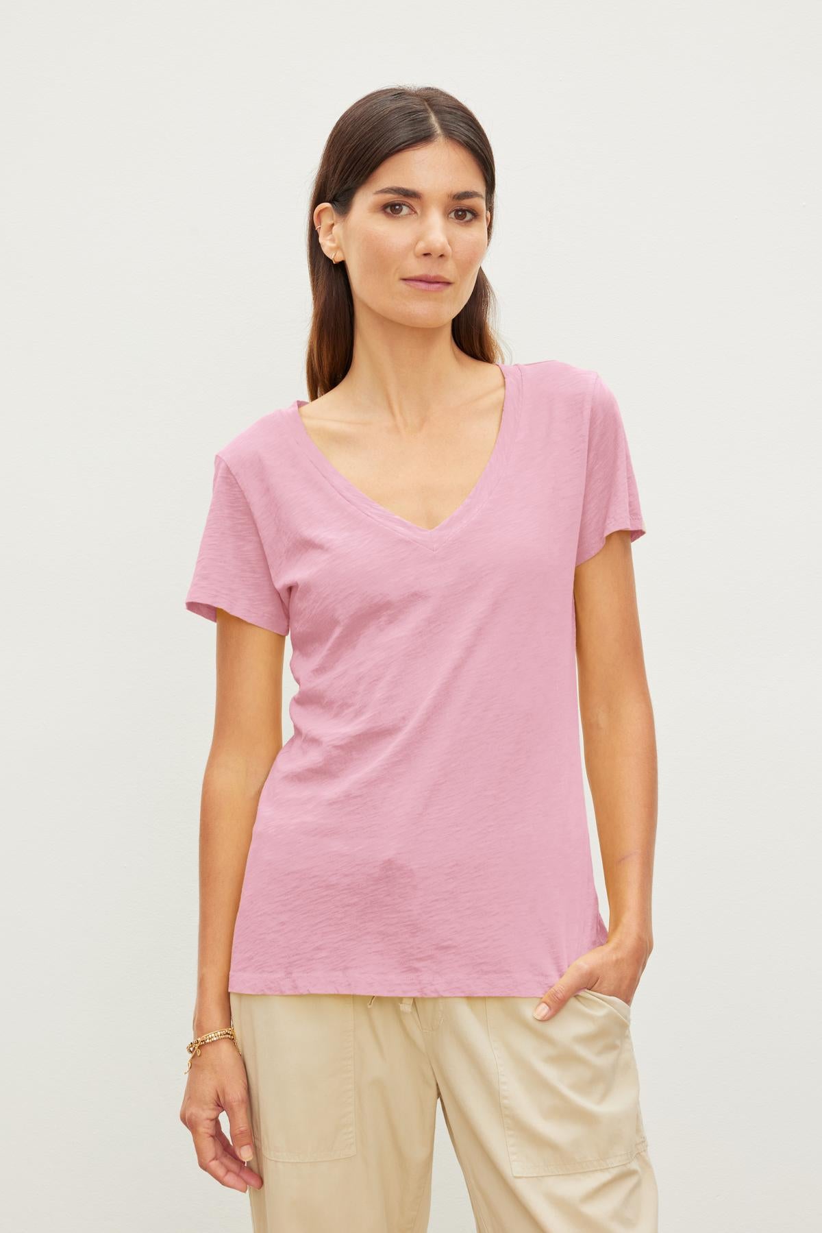 A woman with long dark hair stands against a plain background wearing a pink LILITH TEE by Velvet by Graham & Spencer and beige pants.-37241138774209