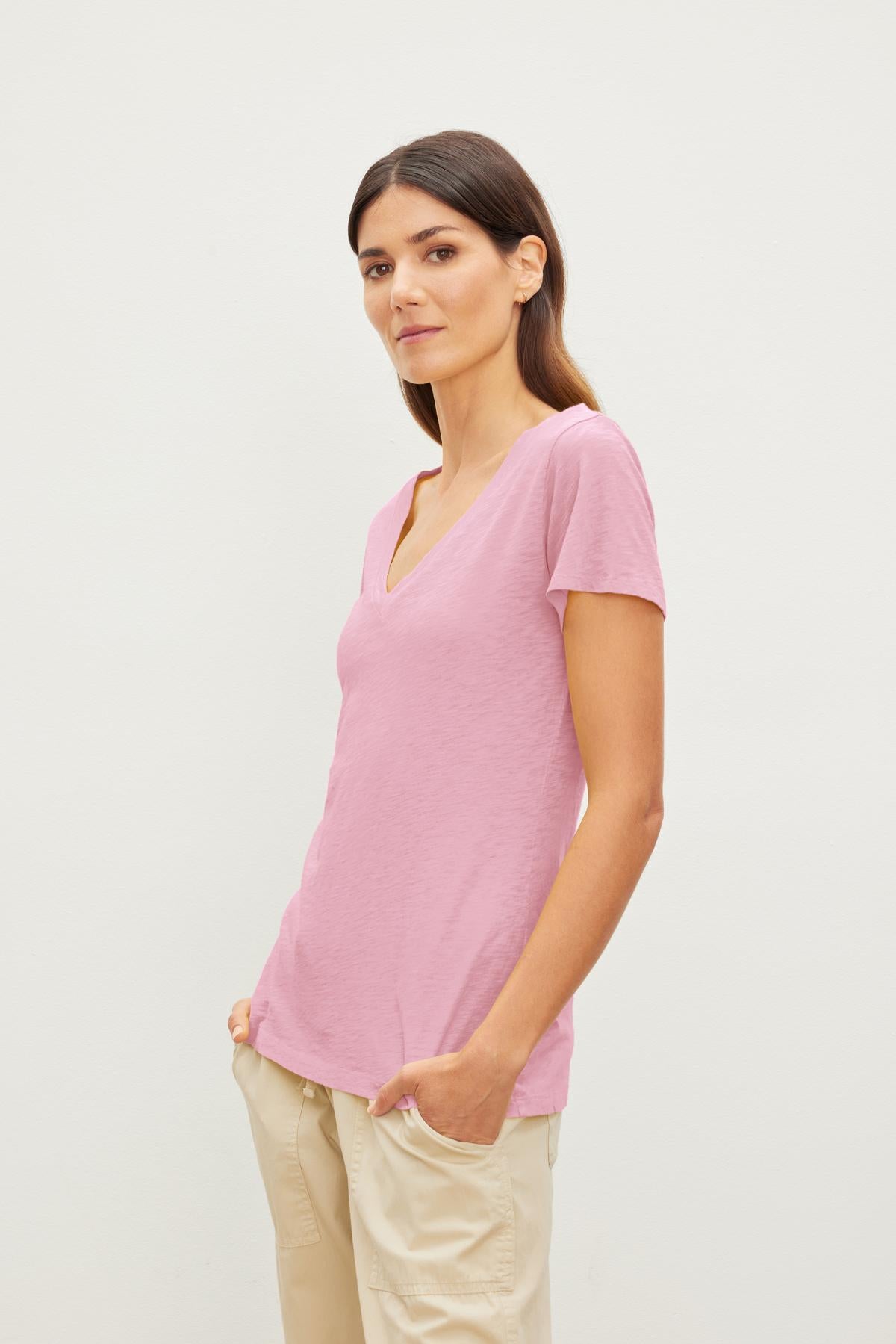   A person wearing a light pink Velvet by Graham & Spencer LILITH TEE and beige pants stands against a plain white background with hands in pockets. 