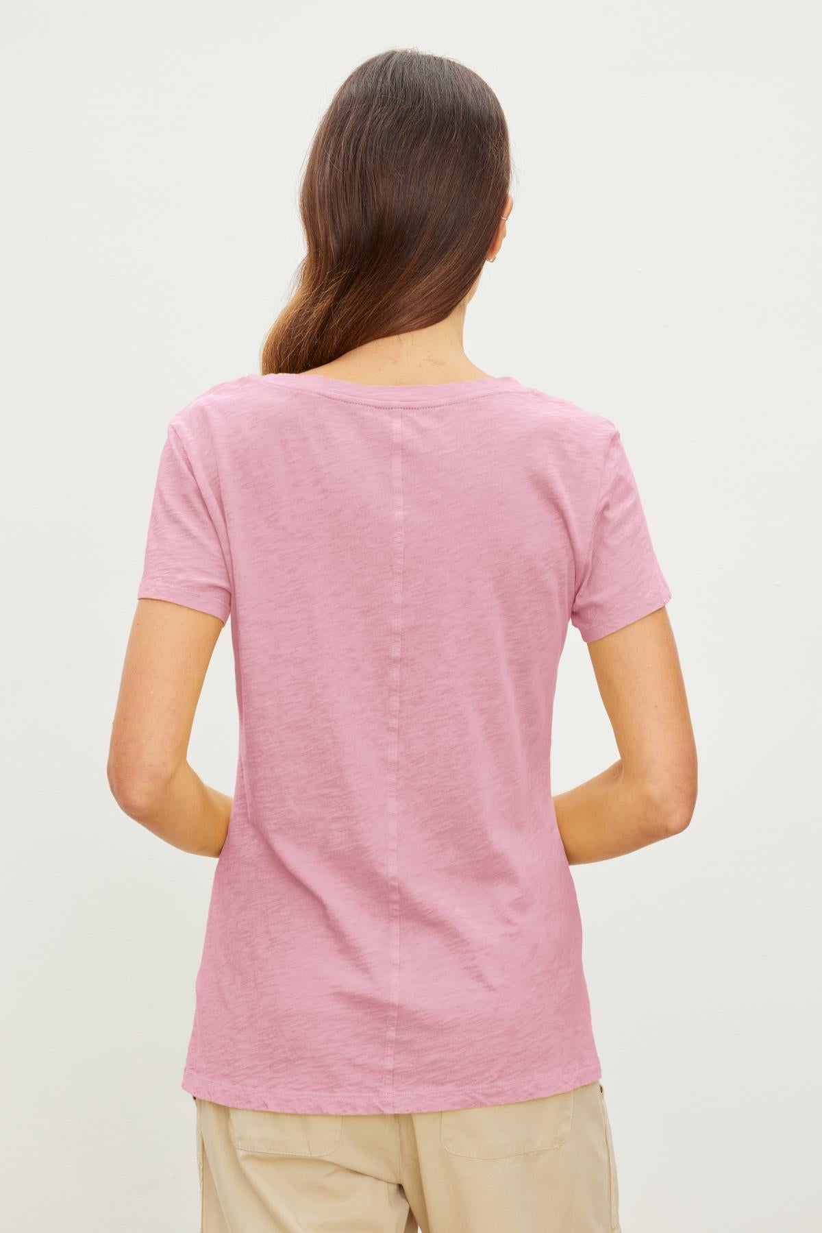   A person with long brown hair stands facing away, wearing a pink Velvet by Graham & Spencer LILITH TEE known for its luxurious softness and beige pants against a plain white background. 