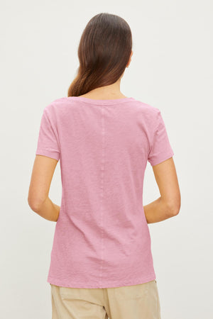 A person with long brown hair stands facing away, wearing a pink Velvet by Graham & Spencer LILITH TEE known for its luxurious softness and beige pants against a plain white background.