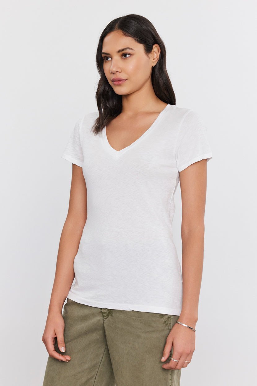 A person stands wearing a white LILITH TEE by Velvet by Graham & Spencer in luxurious cotton slub and olive green pants, looking to the side. The background is plain white.