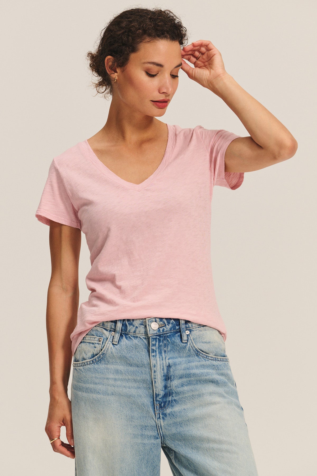 A person wearing the LILITH TEE by Velvet by Graham & Spencer in light pink with light blue jeans stands against a plain background, looking down with one hand touching their head.