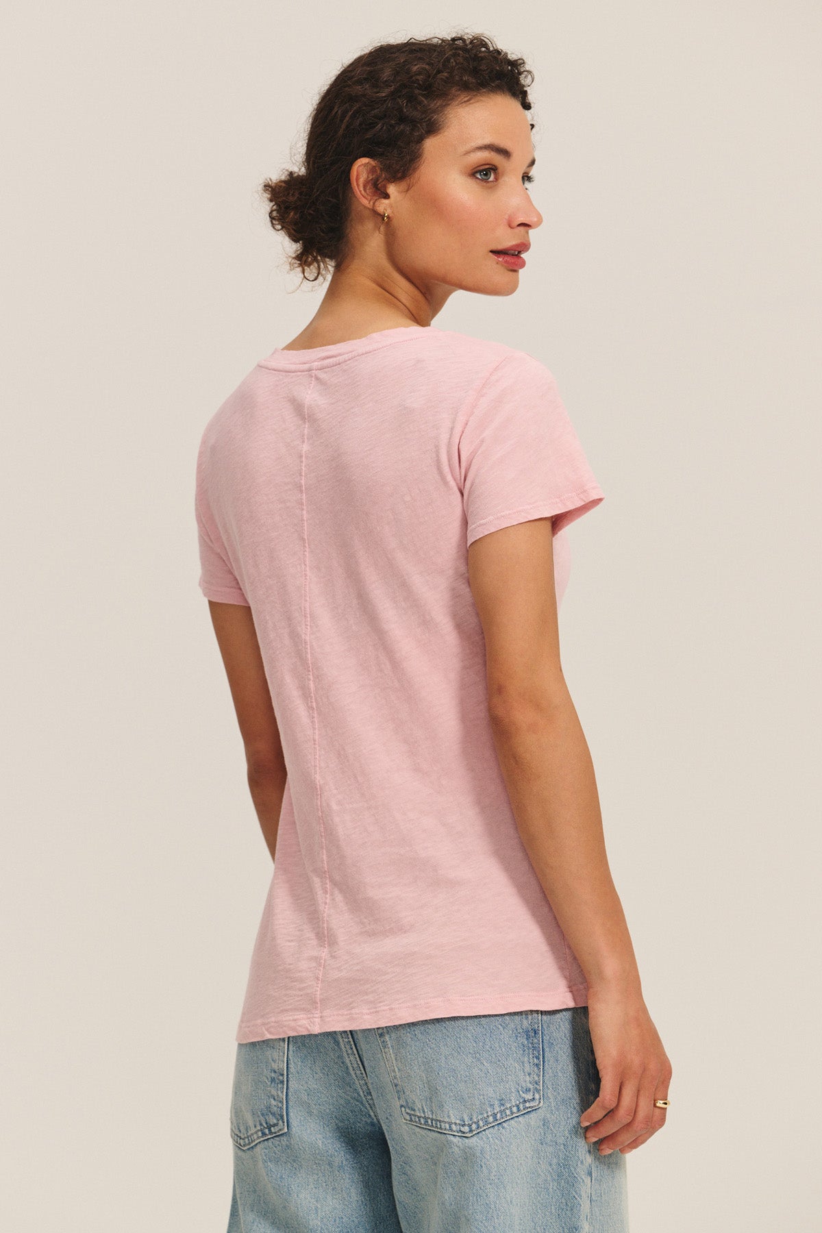 A woman faces away, displaying the back of the Velvet by Graham & Spencer LILITH TEE in light pink short sleeves with light blue jeans against a plain background.