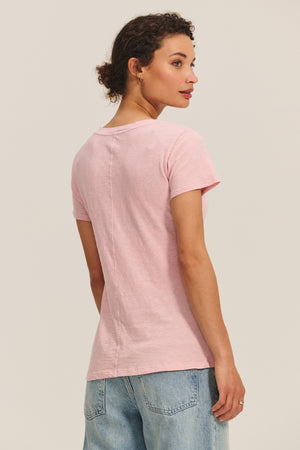 A woman faces away, displaying the back of the Velvet by Graham & Spencer LILITH TEE in light pink short sleeves with light blue jeans against a plain background.
