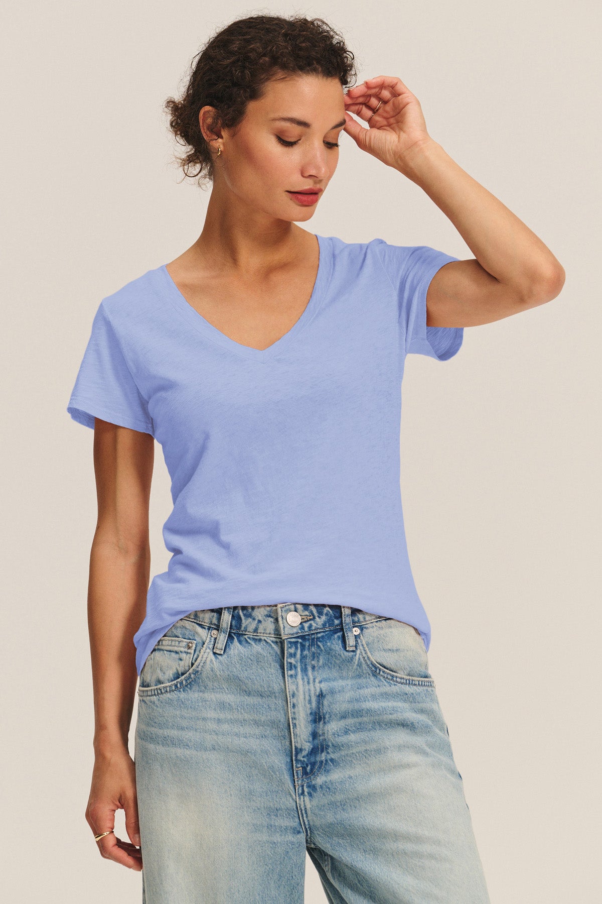A person wears the LILITH TEE by Velvet by Graham & Spencer—a light blue V-neck with a flattering fit—paired with light-wash jeans, standing against a plain background and looking down with one hand touching their head.