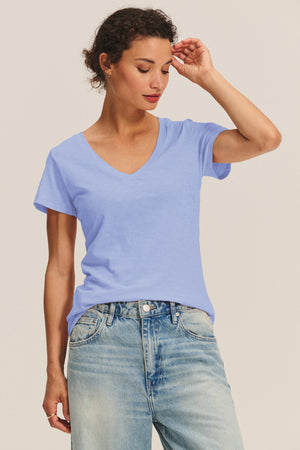 A person wears the LILITH TEE by Velvet by Graham & Spencer—a light blue V-neck with a flattering fit—paired with light-wash jeans, standing against a plain background and looking down with one hand touching their head.