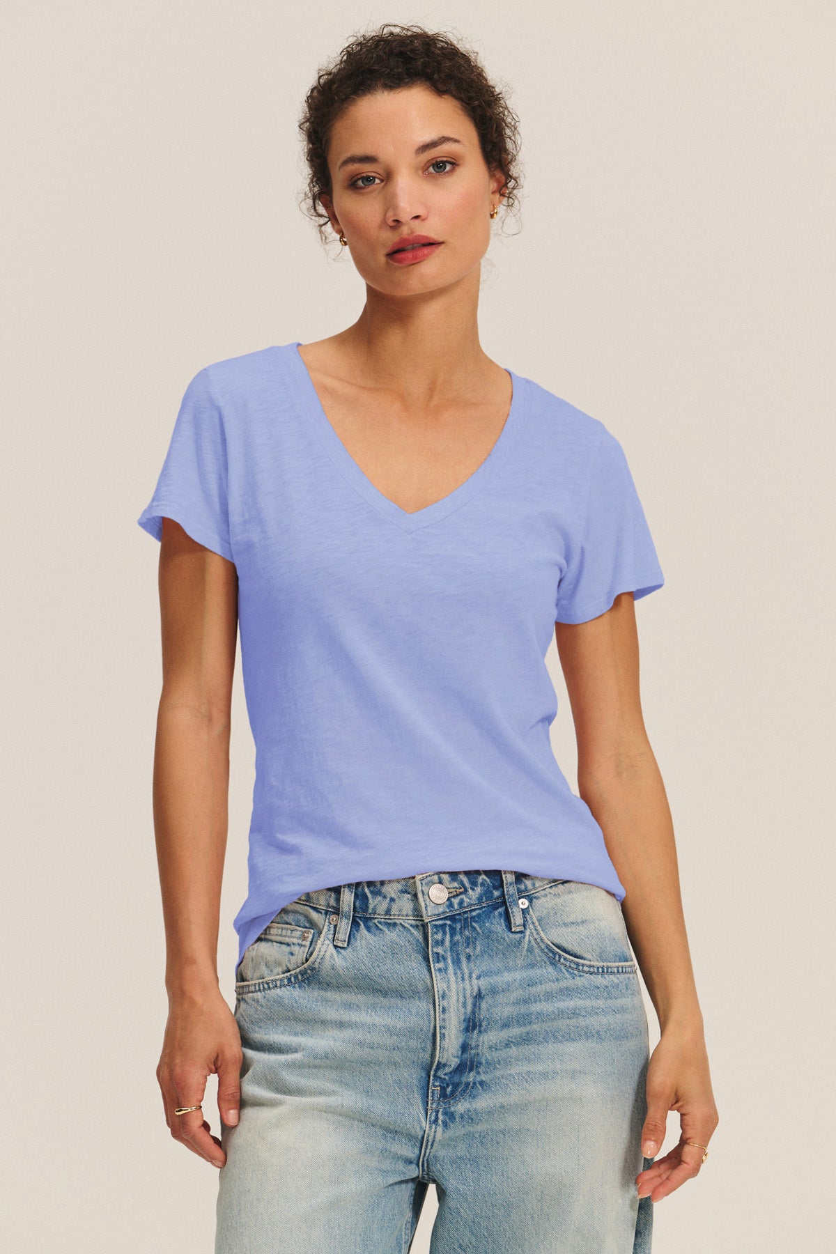 A person stands against a plain background, wearing the LILITH TEE by Velvet by Graham & Spencer in light blue cotton slub with a V-neck, paired with light-wash jeans, looking directly at the camera.