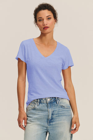 A person stands against a plain background, wearing the LILITH TEE by Velvet by Graham & Spencer in light blue cotton slub with a V-neck, paired with light-wash jeans, looking directly at the camera.
