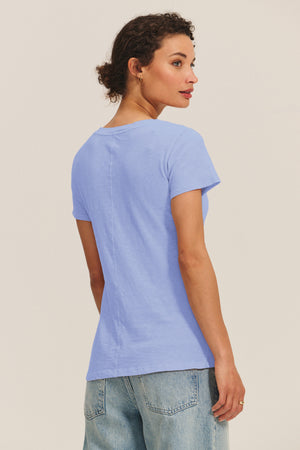 A woman stands against a plain background, looking over her shoulder while wearing the LILITH TEE by Velvet by Graham & Spencer in light blue cotton slub and light wash jeans.