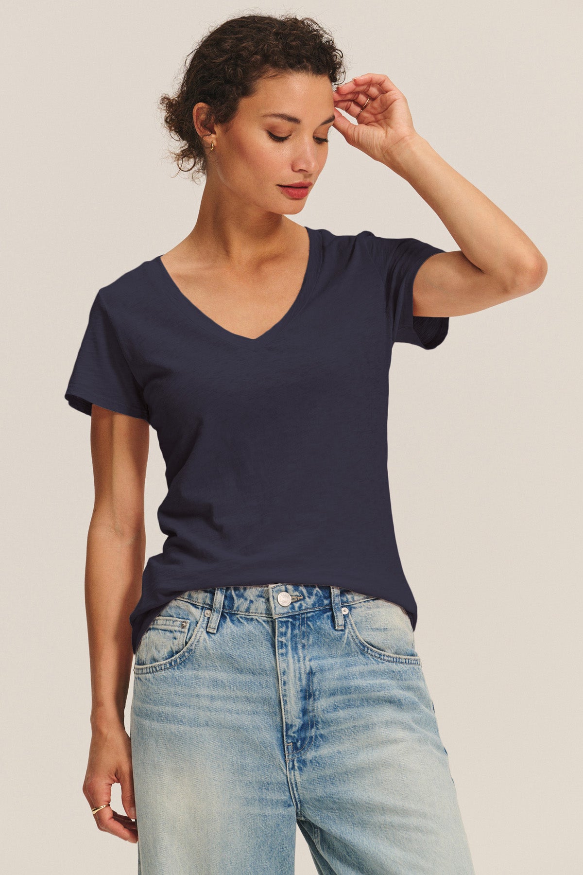 A woman wears the LILITH TEE by Velvet by Graham & Spencer—a plain navy cotton slub V-neck—paired with light blue jeans, standing against a neutral background with her right hand touching her head.-40946704777409