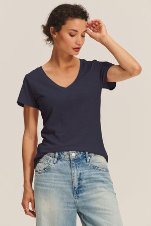 A woman wears the LILITH TEE by Velvet by Graham & Spencer—a plain navy cotton slub V-neck—paired with light blue jeans, standing against a neutral background with her right hand touching her head.