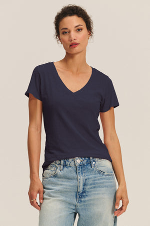 A woman models the LILITH TEE by Velvet by Graham & Spencer—a navy blue cotton slub V-neck—paired with light blue jeans, standing against a plain, light background to highlight the tee’s flattering fit.