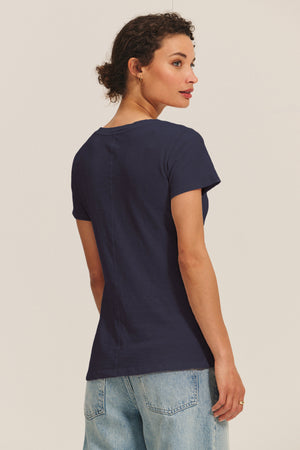 A woman faces away, wearing the LILITH TEE by Velvet by Graham & Spencer—a navy V-neck with a flattering fit—paired with light blue jeans against a plain light background.
