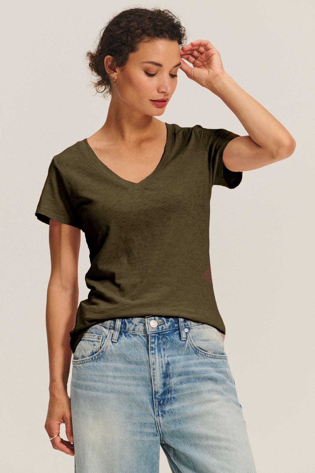 A woman in the LILITH TEE by Velvet by Graham & Spencer, a dark olive women's V-neck in soft cotton slub, pairs it with light blue jeans and stands against a plain background, looking down as she touches her head.