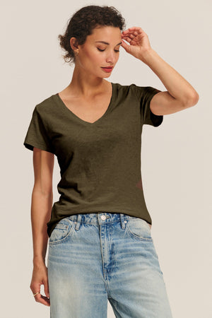 A woman in the LILITH TEE by Velvet by Graham & Spencer, a dark olive women's V-neck in soft cotton slub, pairs it with light blue jeans and stands against a plain background, looking down as she touches her head.