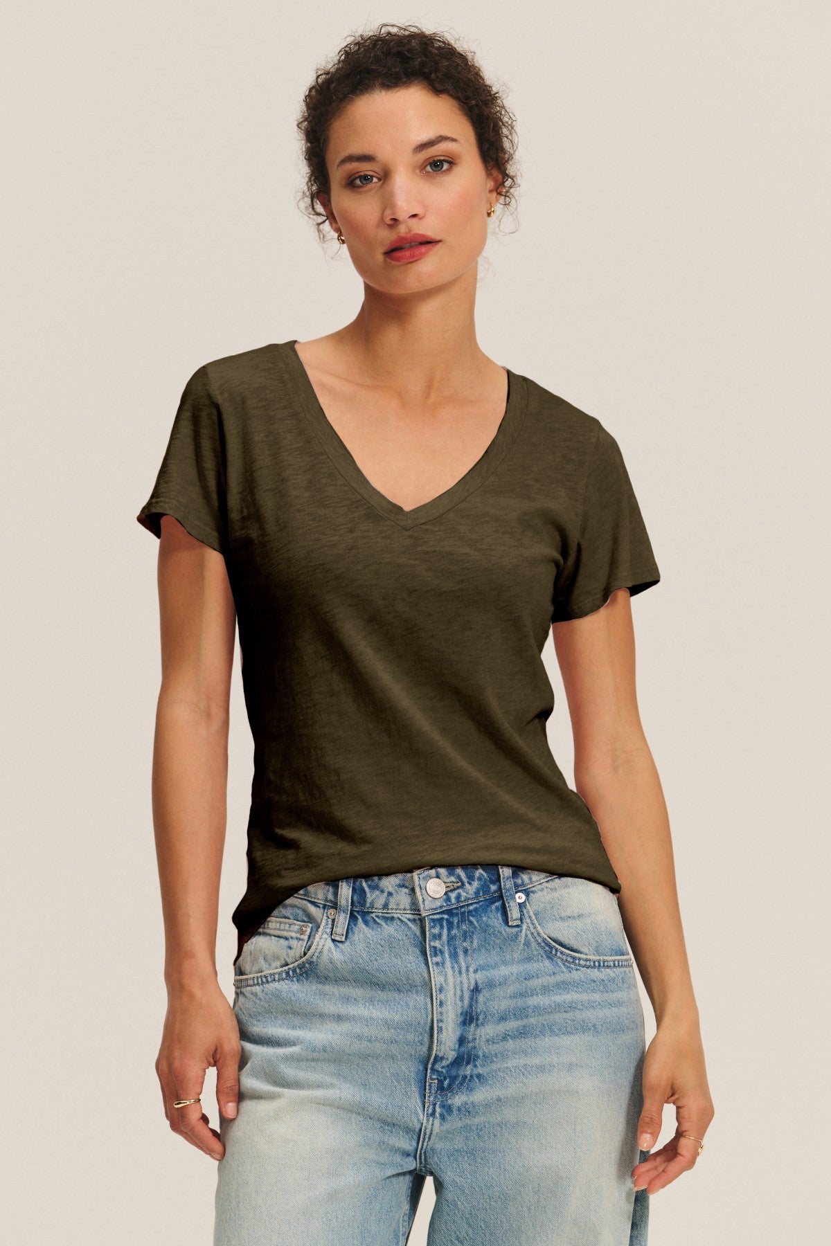 A person stands against a plain, light background wearing the LILITH TEE by Velvet by Graham & Spencer in dark olive cotton slub and paired with light blue jeans.