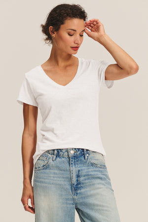A person wearing the LILITH TEE by Velvet by Graham & Spencer—a flattering cotton slub V-neck—paired with light blue jeans stands against a neutral background, looking down with one hand raised to their head.