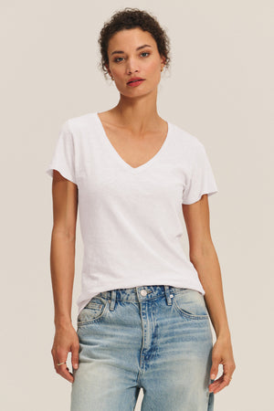 A woman wears the LILITH TEE by Velvet by Graham & Spencer—a flattering, plain white V-neck—paired with light blue jeans, standing against a neutral background.