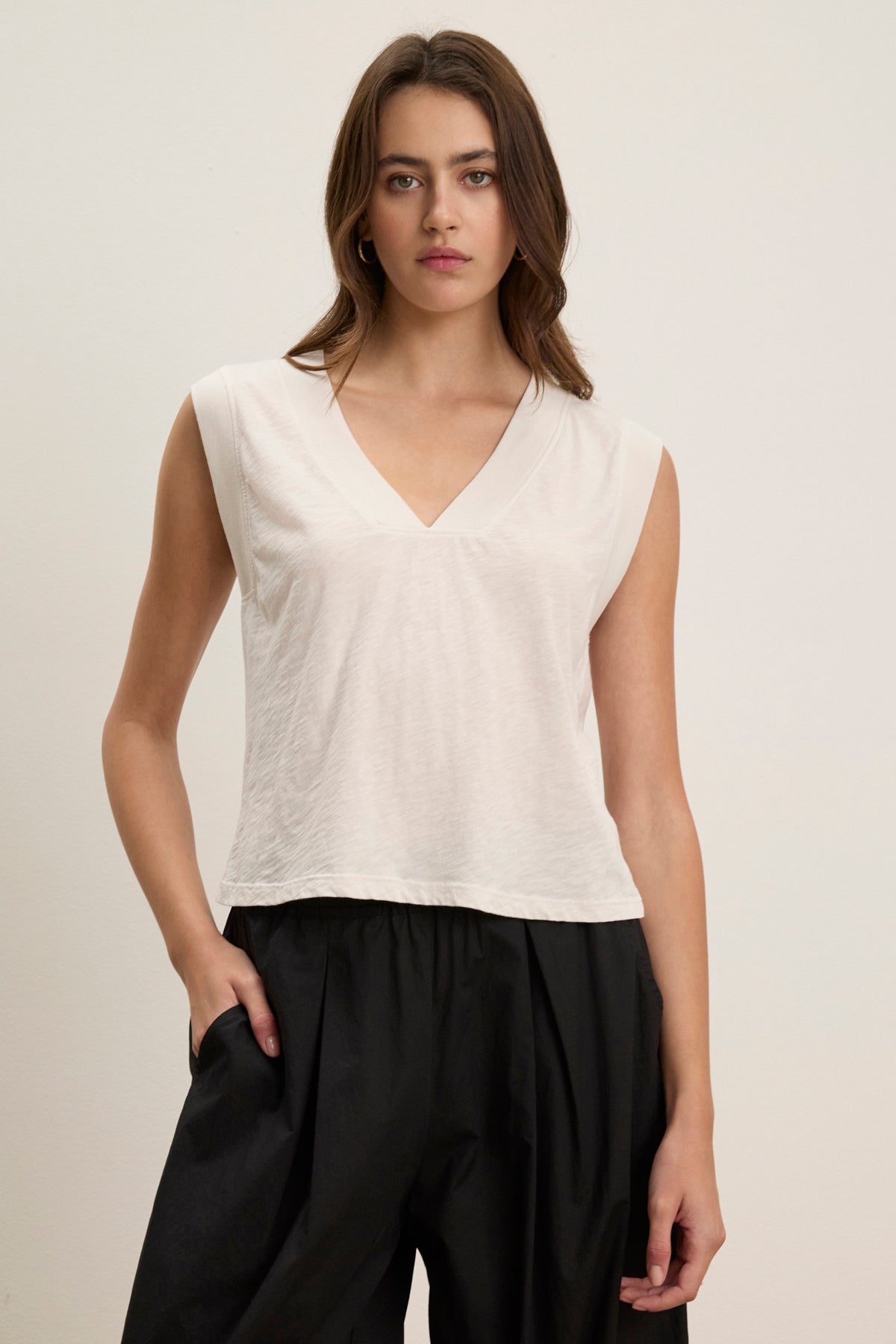 A woman wears the Velvet by Graham & Spencer MARIA TEE, a sleeveless white V-neck top in lightweight breathable textured cotton slub, paired with loose black pants, standing against a plain light-colored background.