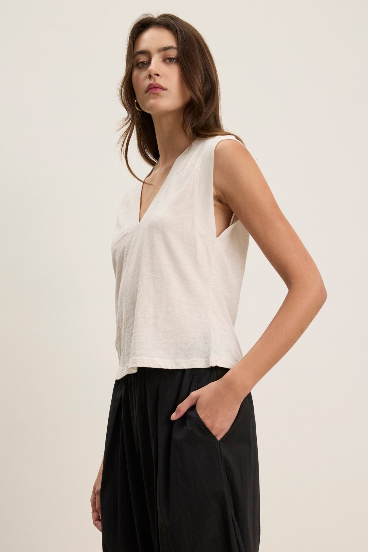 A woman stands against a plain background wearing the MARIA TEE by Velvet by Graham & Spencer, a white sleeveless V-neck top in lightweight, breathable cotton slub, paired with black pants and one hand in her pocket.-39120069230785