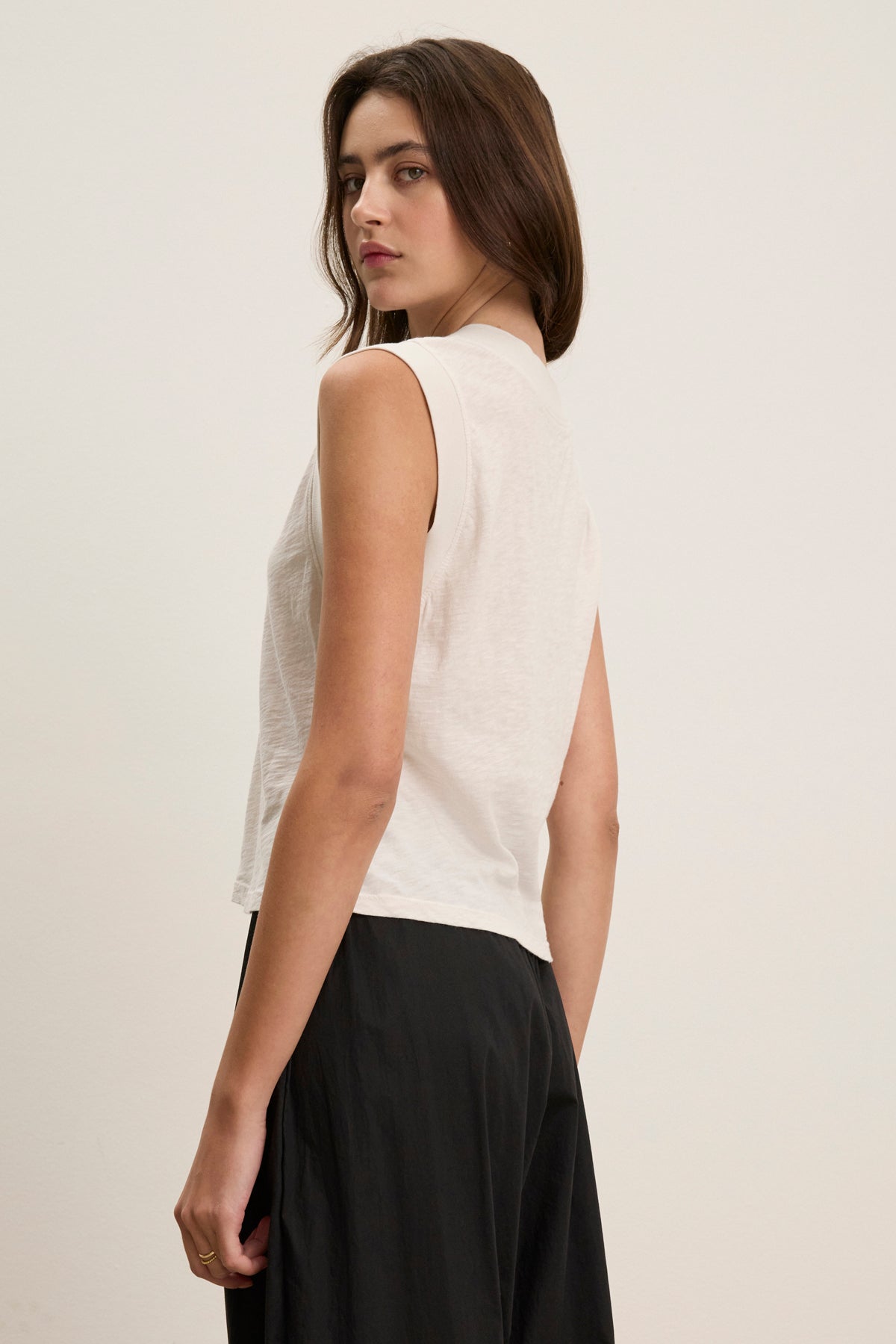 A woman with long brown hair wears the MARIA TEE by Velvet by Graham & Spencer—a sleeveless white top in lightweight breathable textured cotton slub—with a black skirt, standing sideways against a plain light background.-39120069165249