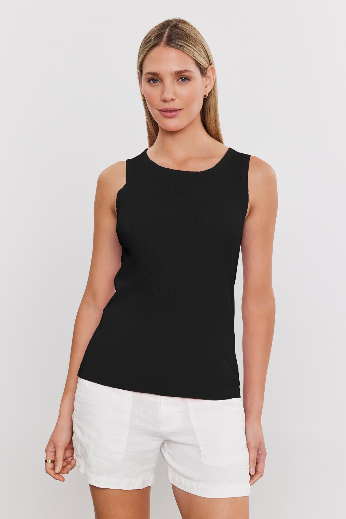   A woman with straight blonde hair, wearing a black sleeveless MAXIE RIBBED TANK TOP by Velvet by Graham & Spencer and white shorts, stands against a plain white background. 