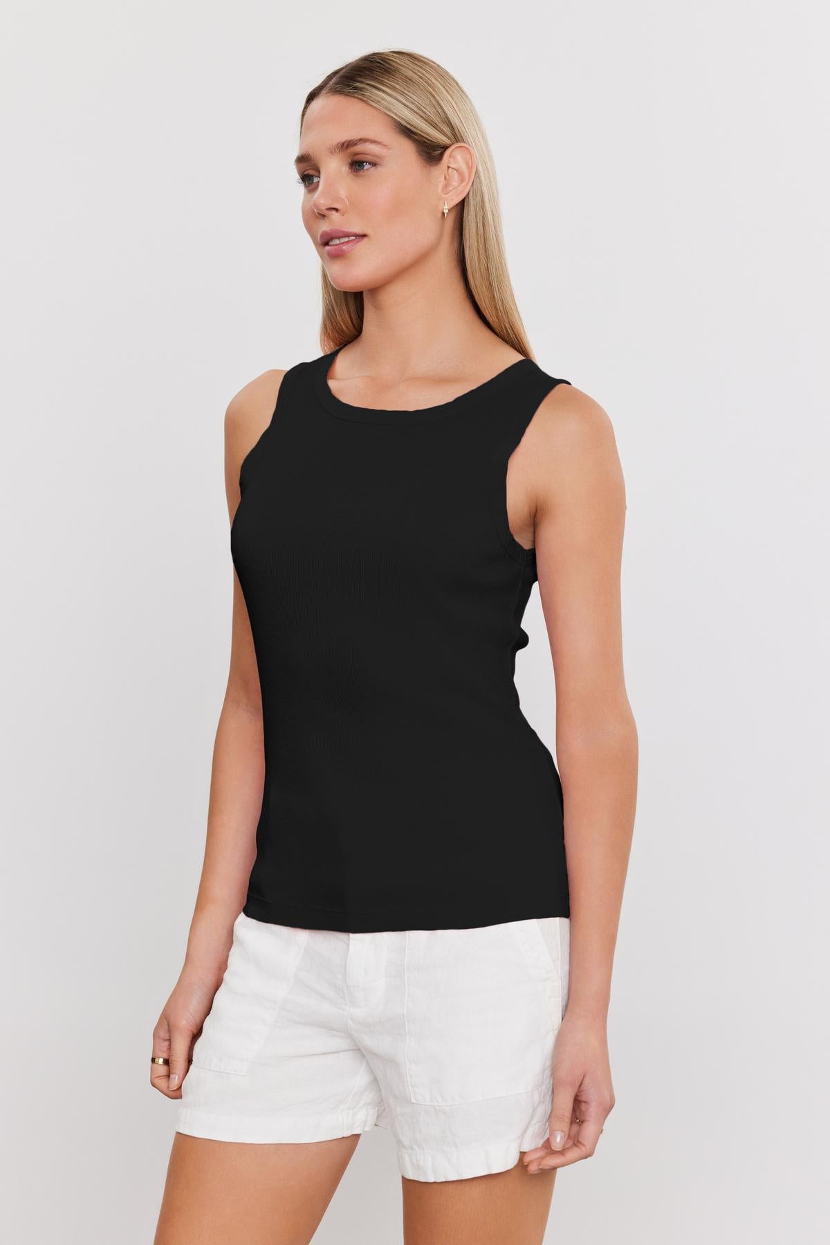 A person with long, straight hair is wearing a sleeveless black top and white shorts, standing against a plain white background. The ribbed knit of the MAXIE RIBBED TANK TOP by Velvet by Graham & Spencer adds a soft textured detail to the outfit.-37241154371777