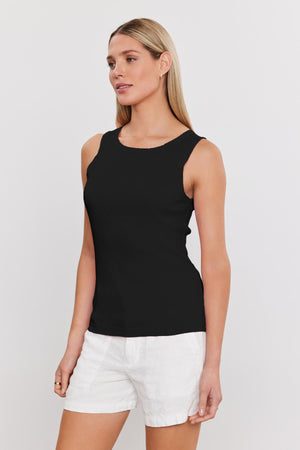 A person with long, straight hair is wearing a sleeveless black top and white shorts, standing against a plain white background. The ribbed knit of the MAXIE RIBBED TANK TOP by Velvet by Graham & Spencer adds a soft textured detail to the outfit.