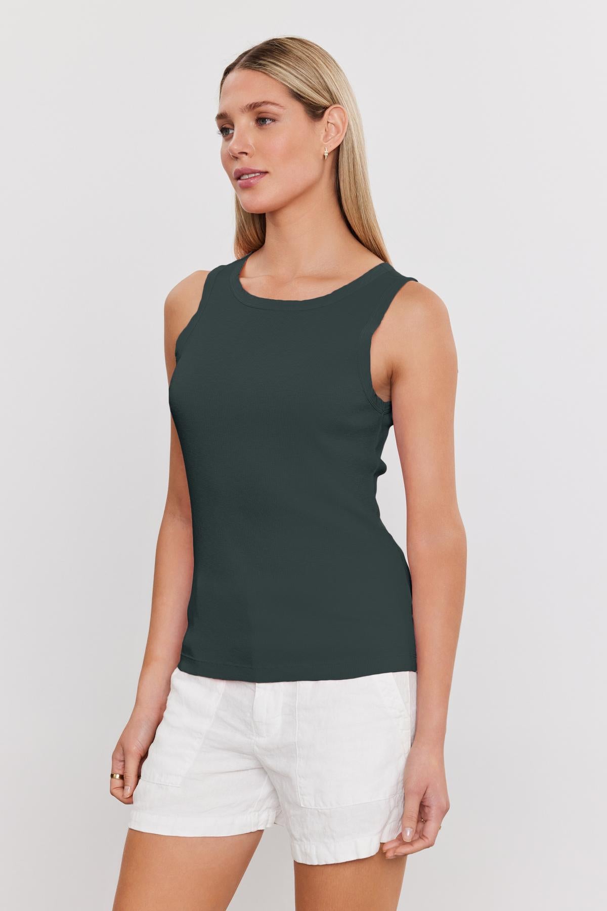 A person with long blonde hair is wearing a dark green MAXIE RIBBED TANK TOP by Velvet by Graham & Spencer and white shorts, standing sideways against a plain white background. The slub knit of the top adds a soft textured detail to their look.-37241154470081