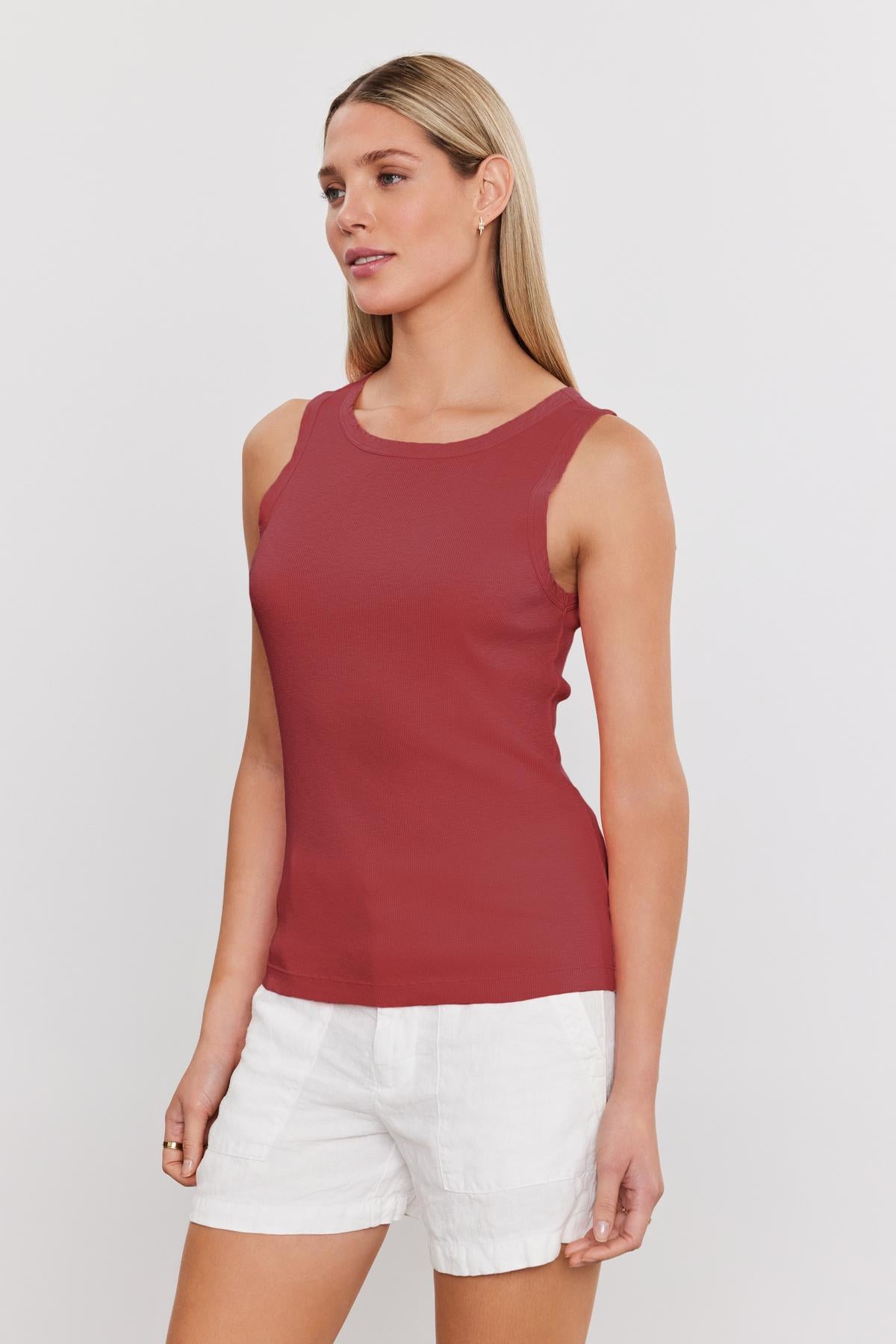 A person wearing a sleeveless red top and white shorts is standing against a plain background, showcasing the soft textured fabric of MAXIE RIBBED TANK TOP by Velvet by Graham & Spencer.-37241154568385