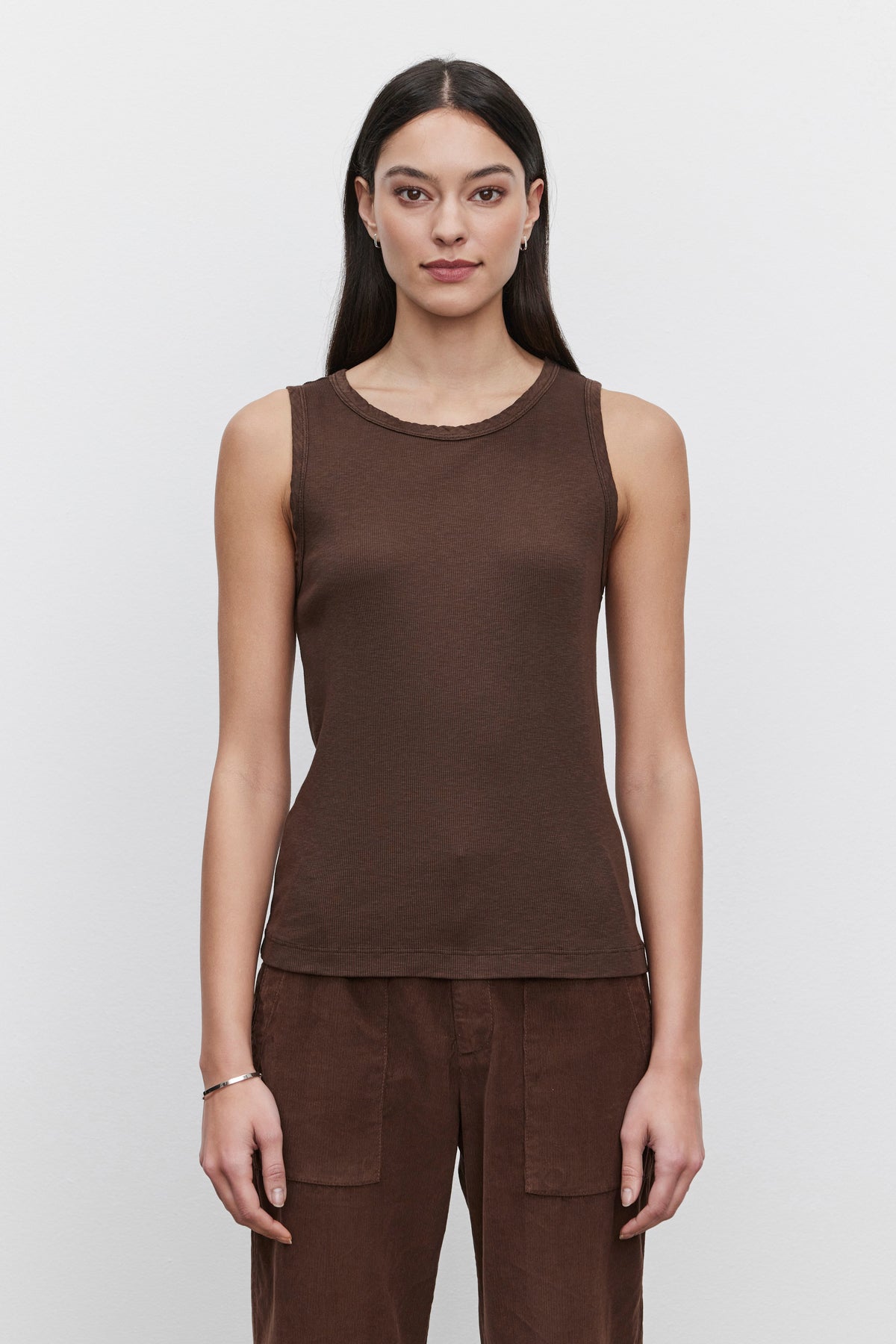   A person with long dark hair, wearing a sleeveless MAXIE RIBBED TANK TOP in brown from Velvet by Graham & Spencer and matching brown pants made from soft textured fabric, stands against a plain white background. 