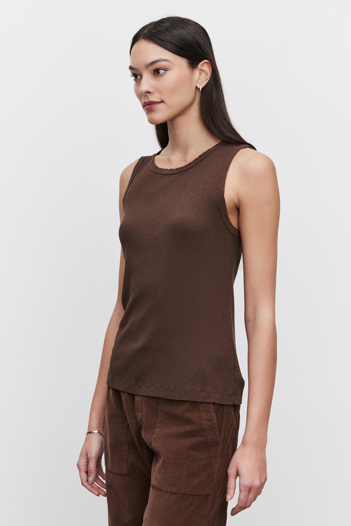   A woman with long dark hair is wearing the MAXIE RIBBED TANK TOP by Velvet by Graham & Spencer, paired with brown pants, standing against a plain white background. Her Maxie tank top features a soft, textured design, creating an effortlessly chic look. 