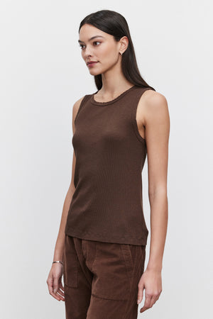 A woman with long dark hair is wearing the MAXIE RIBBED TANK TOP by Velvet by Graham & Spencer, paired with brown pants, standing against a plain white background. Her Maxie tank top features a soft, textured design, creating an effortlessly chic look.