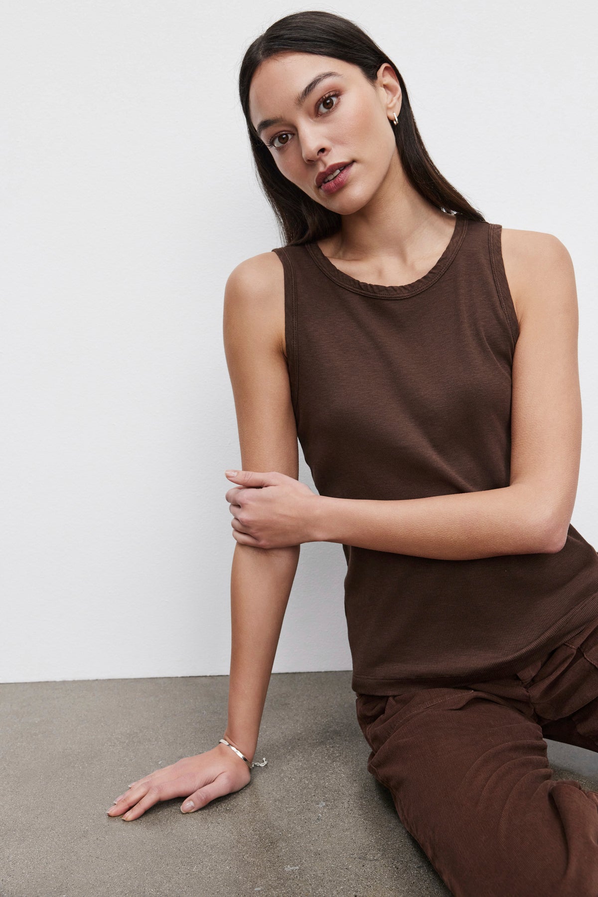 A person with long dark hair, wearing a MAXIE RIBBED TANK TOP by Velvet by Graham & Spencer and soft-textured brown pants, sits on the floor against a plain white wall with one arm resting on their knee.-37665634091201