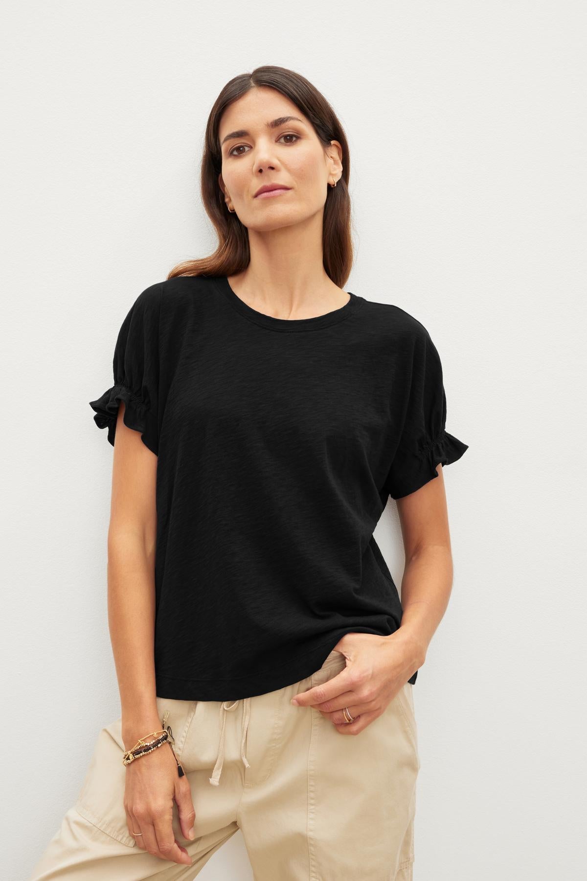   A woman in a black MIMI CREW NECK TEE by Velvet by Graham & Spencer and beige pants stands against a plain background. One hand is in her pocket, and the other rests by her side, showcasing the relaxed fit of her outfit. 