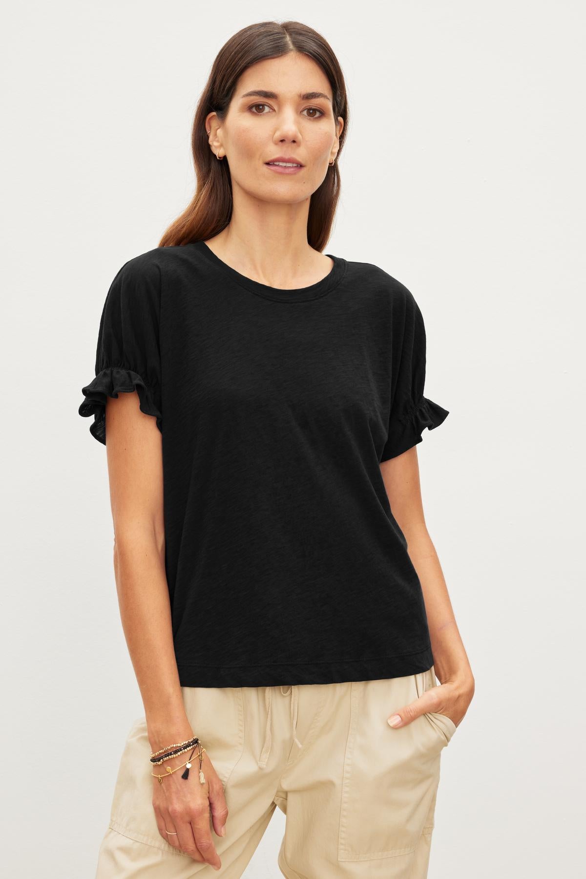   A woman wearing a black MIMI CREW NECK TEE by Velvet by Graham & Spencer with ruffled sleeves and beige pants stands against a plain white background. Her long brown hair cascades down as she poses with one hand in her pocket, showcasing the relaxed fit of her stylish outfit. 