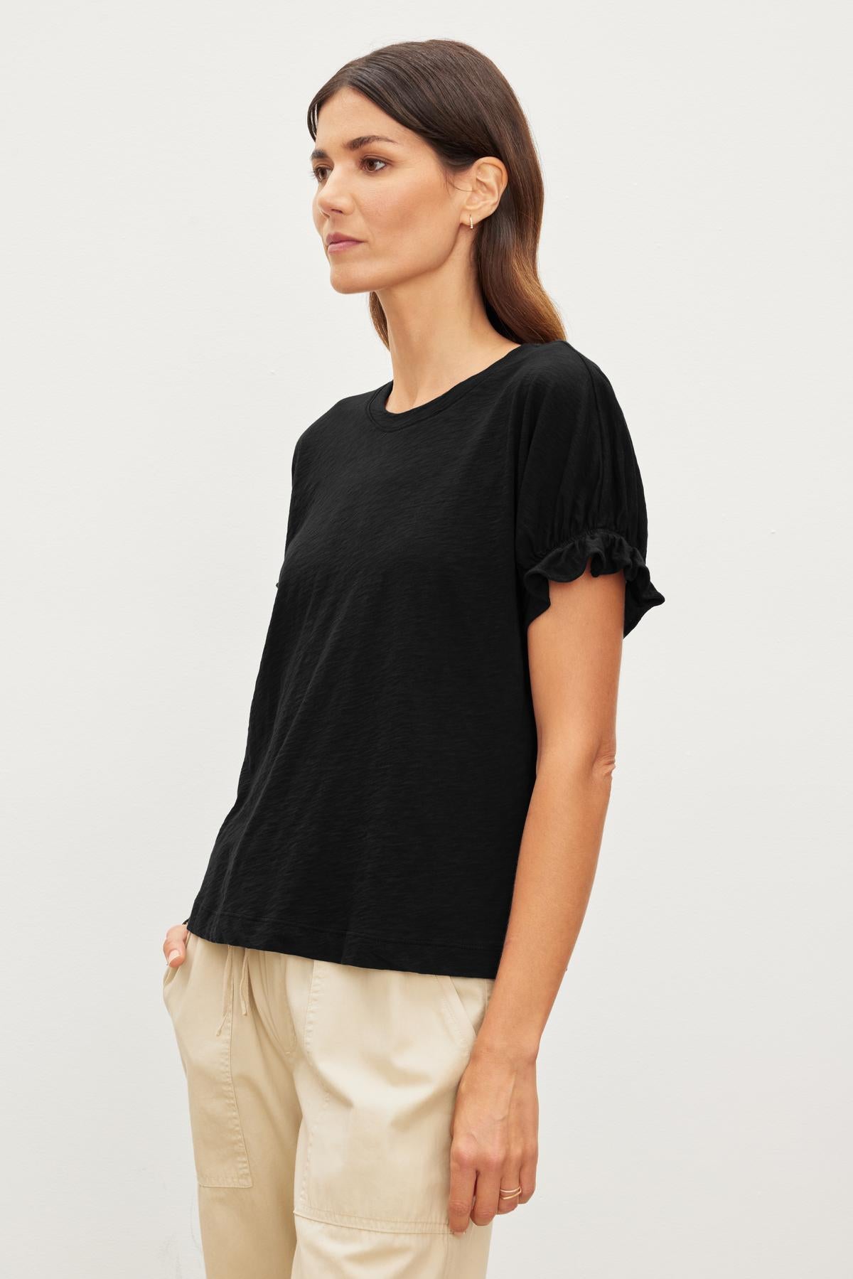   A woman with long brown hair is wearing a black MIMI CREW NECK TEE by Velvet by Graham & Spencer and beige pants. She is standing sideways against a plain white background. 