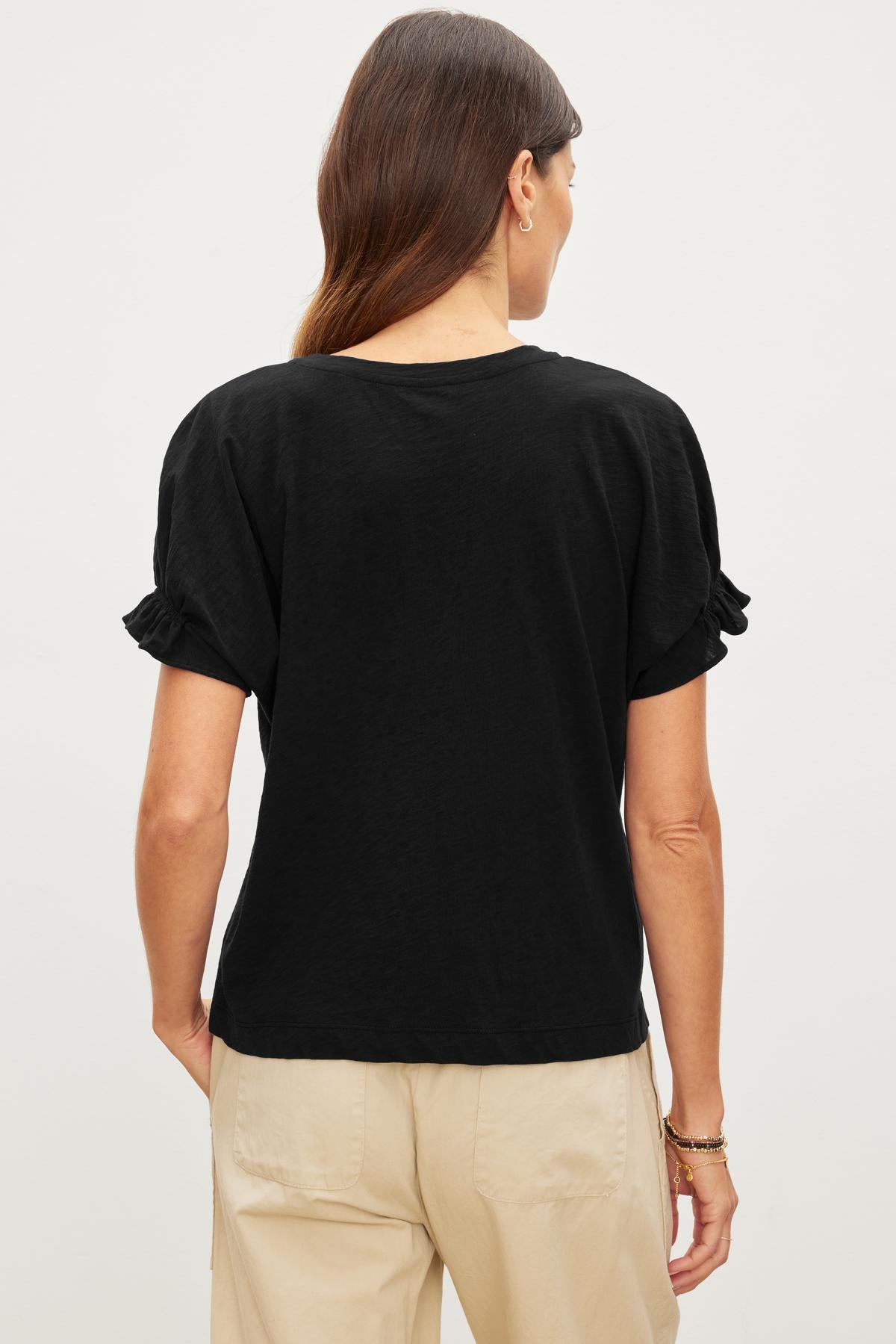   A person stands with their back to the camera, wearing a black MIMI CREW NECK TEE by Velvet by Graham & Spencer and beige pants in a relaxed fit. 